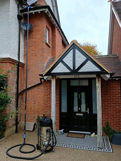 Red brick house entrance with black timber-framed gable porch, black front door, and coiled hose on ground