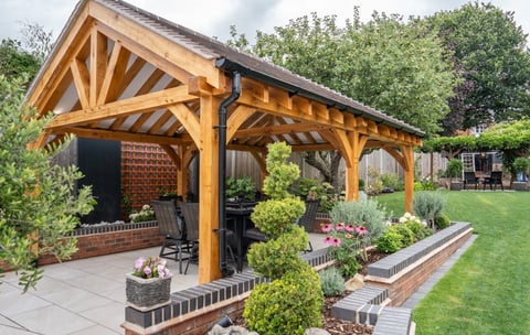 Wooden pergola with timber framing attached to brick house, featuring seating area and landscaped garden with topiary plants