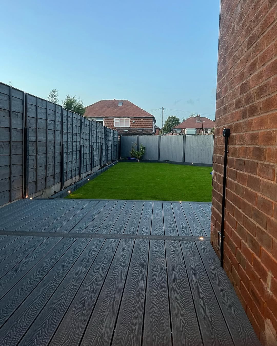 Modern backyard with grey composite decking, artificial green lawn, dark fencing, brick wall, and residential houses in background under clear sky