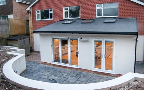 Modern white single-story extension with orange trim and glass doors attached to a brick house, featuring a curved patio area.