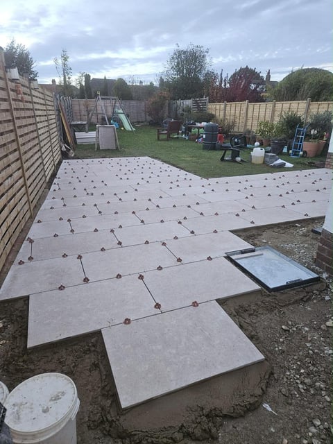 Backyard patio installation with grey paving stones laid out on soil, secured with brown fasteners, surrounded by fenced garden and landscaping