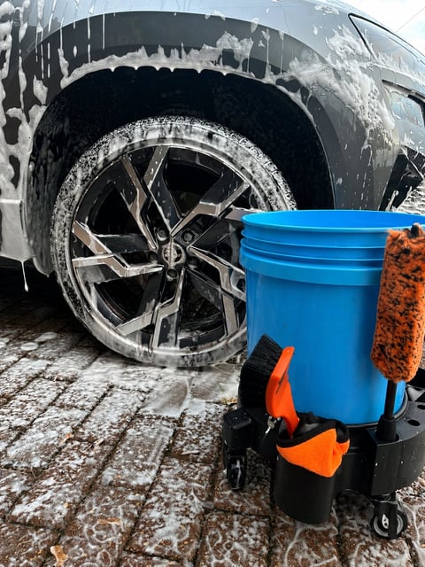 Car wheel being washed with blue bucket containing soapy water and orange sponge on snowy surface
