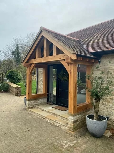 Modern oak timber frame entrance porch with black door and glass panels attached to stone cottage with slate roof