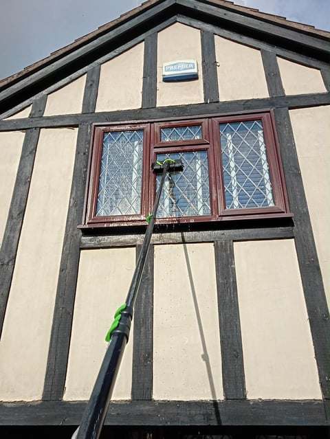 Historic half-timbered building with cream and dark timber framing, red leaded windows, and a green cleaning pole in foreground