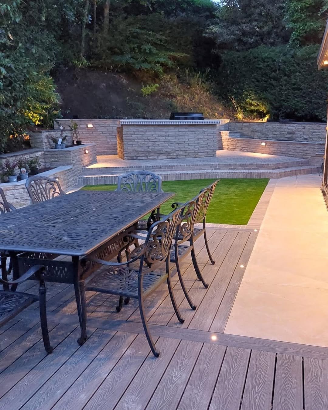 Backyard patio with metal dining table, chairs, manicured lawn, stone retaining wall with built-in seating, and evening landscape lighting