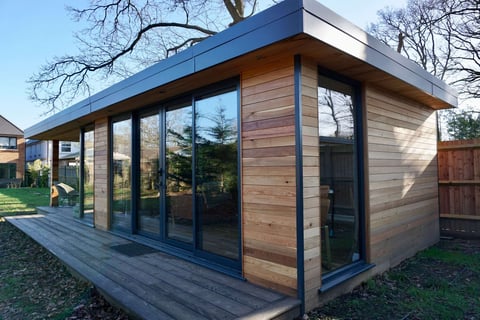 Modern garden studio with blue roof, wooden siding, large windows, and wooden deck surrounded by bare trees