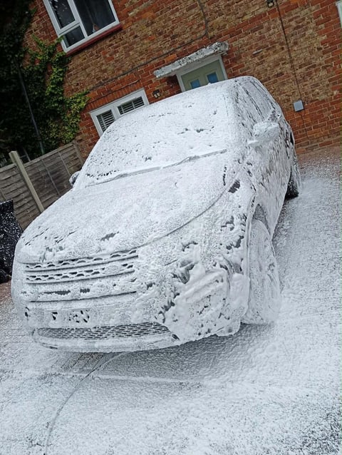 Car covered in white foam or soap suds parked on a driveway in front of a brick house