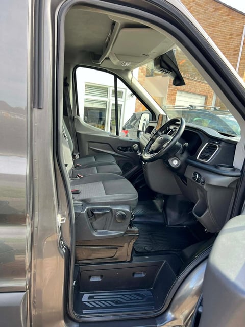 Interior view of a utility vehicle with open drivers door, steering wheel, dashboard, and gray upholstered seats, parked outside a brick building